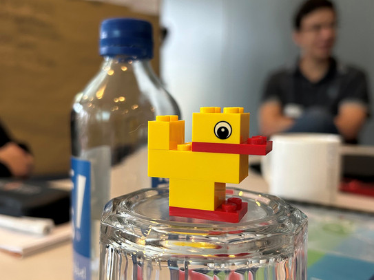 Foto: Eine aus Bausteinen zusammengesetzte Ente auf einem umgedrehten Wasserglas, daneben eine Flasche, im Hintergrund eine Pinnwand und eine sitzende Person