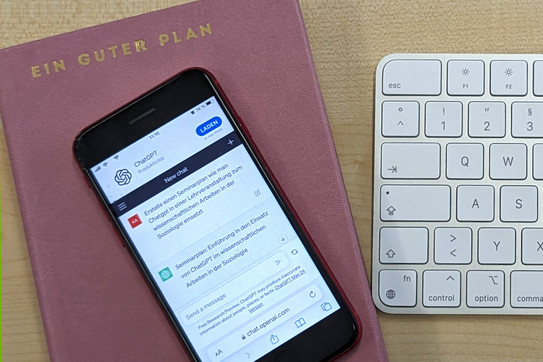 Foto: Ein Mobiltelefon, auf dem ChatGPT geöffnet ist, liegt auf einem Terminplaner, der mit dem Schriftzug "Ein guter Plan" betitelt ist. Rechts daneben liegt eine weiße Rechnertastatur