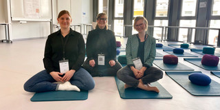 Foto: Joana Eichhorn, Dr. Katrin Stolz und Dr. Julia Mergner sitzen im Schneidersitz auf Yogamatten in einem Tagungsraum