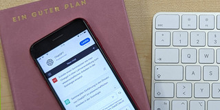 Foto: Ein Mobiltelefon, auf dem ChatGPT geöffnet ist, liegt auf einem Terminplaner, der mit dem Schriftzug "Ein guter Plan" betitelt ist. Rechts daneben liegt eine weiße Rechnertastatur