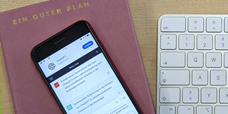 Foto: Ein Mobiltelefon, auf dem ChatGPT geöffnet ist, liegt auf einem Terminplaner, der mit dem Schriftzug "Ein guter Plan" betitelt ist. Rechts daneben liegt eine weiße Rechnertastatur