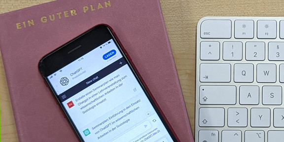 Foto: Ein Mobiltelefon, auf dem ChatGPT geöffnet ist, liegt auf einem Terminplaner, der mit dem Schriftzug "Ein guter Plan" betitelt ist. Rechts daneben liegt eine weiße Rechnertastatur