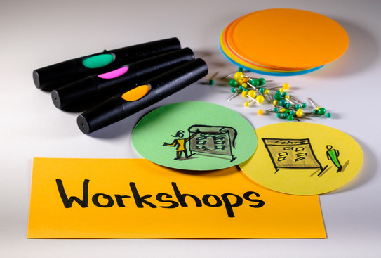 Foto mit Moderationsmaterialien: Im Vordergrund eine rechteckige Metaplankarte mit dem Wort "Workshops", rechts darüber zwei runde Metaplankarten mit gezeichneten Workshopszenen, darüber Moderationsstifte, Pinnadeln und ein Stapel runde Metaplankarten