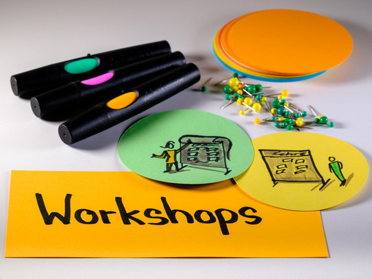 Foto mit Moderationsmaterialien: Im Vordergrund eine rechteckige Metaplankarte mit dem Wort "Workshops", rechts darüber zwei runde Metaplankarten mit gezeichneten Workshopszenen, darüber Moderationsstifte, Pinnadeln und ein Stapel runde Metaplankarten