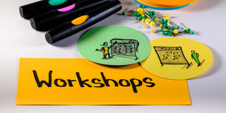 Foto mit Moderationsmaterialien: Im Vordergrund eine rechteckige Metaplankarte mit dem Wort "Workshops", rechts darüber zwei runde Metaplankarten mit gezeichneten Workshopszenen, darüber Moderationsstifte, Pinnadeln und ein Stapel runde Metaplankarten