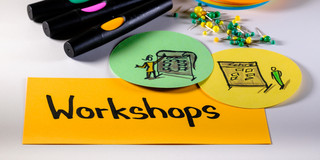 Foto mit Moderationsmaterialien: Im Vordergrund eine rechteckige Metaplankarte mit dem Wort "Workshops", rechts darüber zwei runde Metaplankarten mit gezeichneten Workshopszenen, darüber Moderationsstifte, Pinnadeln und ein Stapel runde Metaplankarten