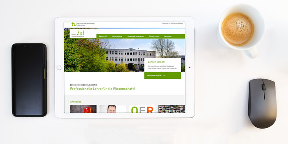 Tablet mit der Webseite des Bereichs Hochschuldidaktik, daneben ein Mobiltelefon, eine Kaffeetasse und eine Computermaus