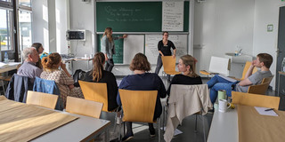 Foto einer Workshopszene: Die Dozentinnen stehen an einer Tafel, auf der Workshopinhalte notiert und Flipchartpapiere angeheftet sind. Die Teilnehmenden sitzen im Stuhlkreis davor und betrachten die Dozierenden und die Tafel.