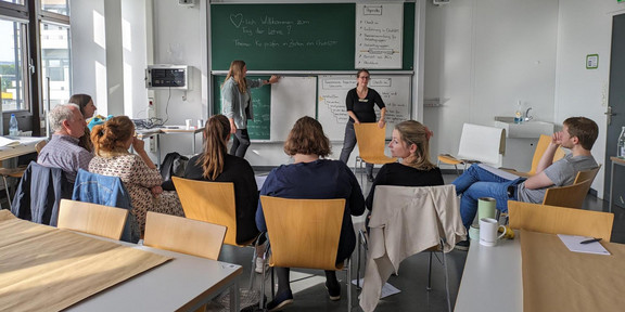 Foto einer Workshopszene: Die Dozentinnen stehen an einer Tafel, auf der Workshopinhalte notiert und Flipchartpapiere angeheftet sind. Die Teilnehmenden sitzen im Stuhlkreis davor und betrachten die Dozierenden und die Tafel.