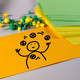 Foto von Moderationsmaterialien: eine orange rechteckige Moderationskarte mit einer Zeichnung von einer jonglierenden Person darauf. Im Hintergrund grüne und gelbe Pinnadeln und Moderationskarten
