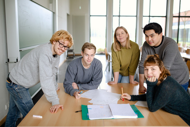 Fünf Studierende im Seminarraum stehen um einen Tisch herum und arbeiten gemeinsam