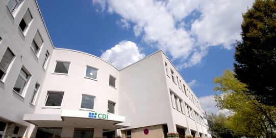 CDI building: The entrance of a white building with two wings from the frog's perspective. In front of it a bicycle stand and right in front of the building trees.