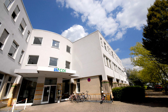 CDI building: The entrance of a white building with two wings from the frog's perspective. In front of it a bicycle stand and right in front of the building trees.